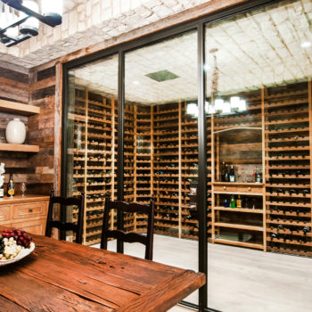 A rustic dining area featuring Euroline Steel Wine Room Doors, with black steel frames and glass panels showcasing an extensive wine collection inside a well-lit room, complemented by a wooden dining table and cozy ambiance.