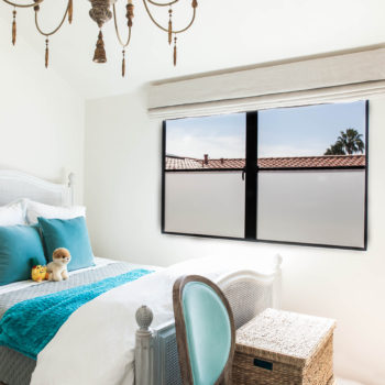 Cozy and inviting bedroom enhanced by Euroline Steel Casement Windows, featuring a large black steel-framed window that provides a clear view of palm trees and a serene outdoor environment. The room is decorated with a comfortable bed, vibrant blue accents, and vintage-style furniture, creating a charming and relaxing space.