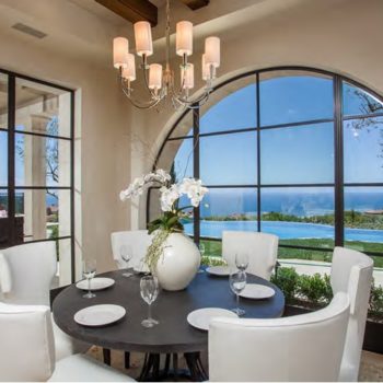 Elegant dining room outfitted with Euroline Steel Curtain Wall Windows, featuring a large arched window that provides stunning ocean views. The interior is tastefully decorated with a round table, white chairs, and a chic chandelier, creating a luxurious dining experience.