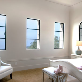 Minimalist and elegant living area illuminated by natural light from Euroline Steel Casement Windows, featuring three vertical black steel-framed windows that provide views of the outdoors. The room is decorated with a white chaise lounge, a plush bench, and soft neutral tones for a serene atmosphere.