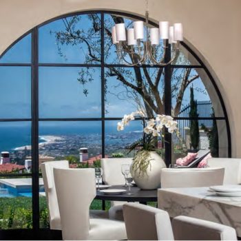 Elegant dining area with Euroline Steel Curtain Wall Windows, showcasing an expansive arched window with black steel frames, providing a panoramic view of the coast, complemented by modern white furniture.