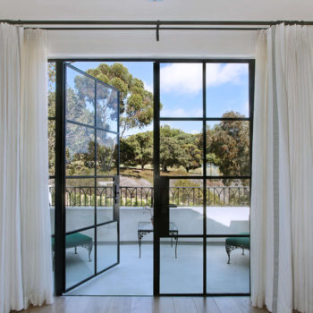 Elegant interior framed by Euroline Steel Swing Doors, featuring floor-to-ceiling black frames that open to a tranquil outdoor patio surrounded by lush greenery. Sheer white curtains add a soft touch to the bold door design, enhancing the room’s bright and airy feel.