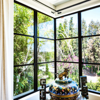 Luxurious corner view from a room featuring Euroline Steel Casement Windows, with large black steel-framed windows that encompass the lush, vibrant garden outside. The interior is decorated with a detailed and colorful ornamental vase, enhancing the connection between the indoor elegance and the natural beauty outside.