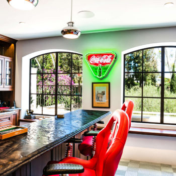 Vibrant home bar area enhanced by Euroline Steel Casement Windows, featuring large windows with black steel frames that offer views of the surrounding green landscape. The bar is uniquely styled with red racing seat bar stools, a neon Coca-Cola sign, and modern pendant lighting, creating a lively and eclectic space.