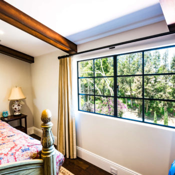 Cozy bedroom featuring Euroline Steel Casement Windows, with a large black steel-framed window offering a picturesque view of lush greenery. The room is tastefully decorated with traditional furnishings, a patterned quilt, and elegant drapery, enhancing the warm and inviting atmosphere.