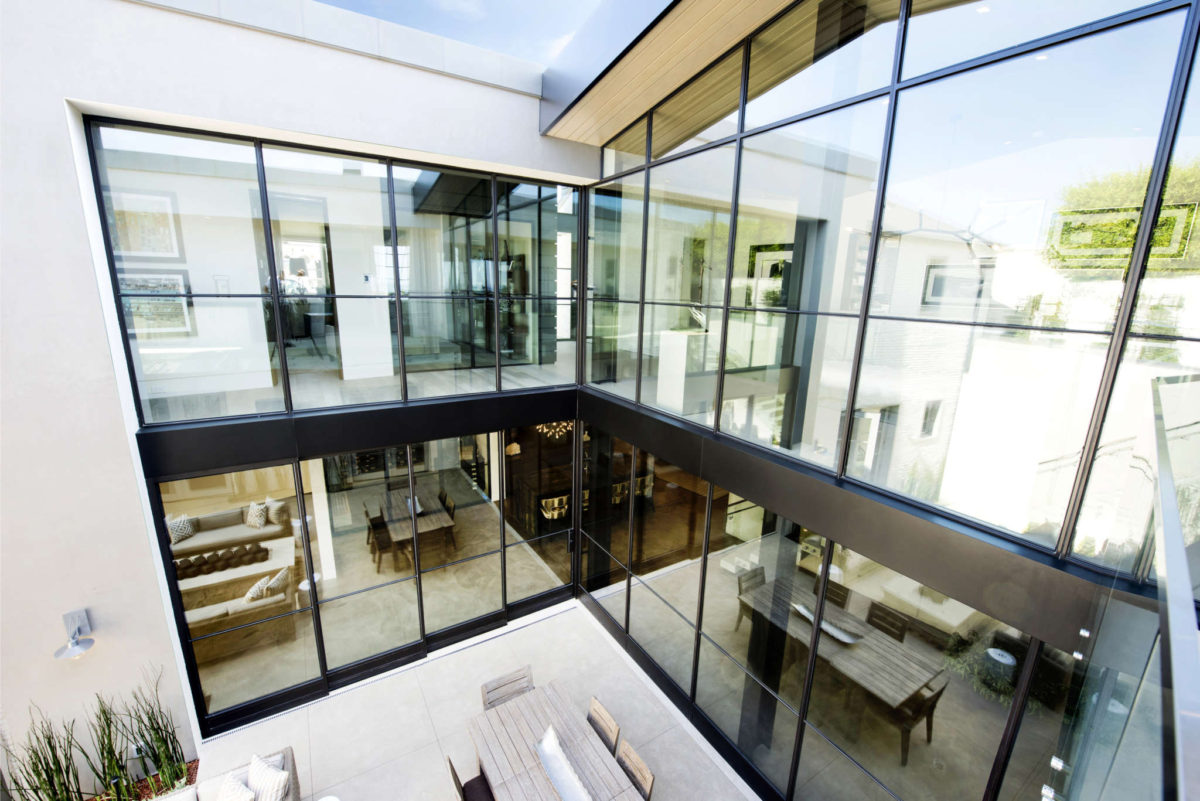 Modern home interior featuring Euroline Steel Curtain Wall Windows, with a two-story glass facade that provides a transparent and continuous view of the stylishly decorated living and dining areas. The black steel frames add a striking contrast to the bright and open architectural design.