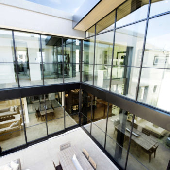 Modern home interior featuring Euroline Steel Curtain Wall Windows, with a two-story glass facade that provides a transparent and continuous view of the stylishly decorated living and dining areas. The black steel frames add a striking contrast to the bright and open architectural design.
