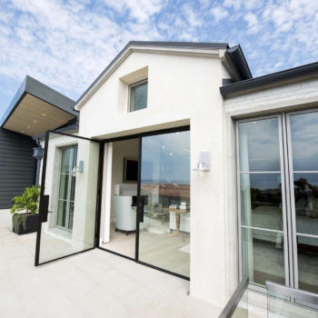 Modern home exterior featuring Euroline Steel Swing Doors and large windows, with clean black frames that contrast beautifully against the home’s white and dark gray siding. The design promotes a seamless indoor-outdoor flow, enhancing the connection to the spacious terrace and scenic surroundings.