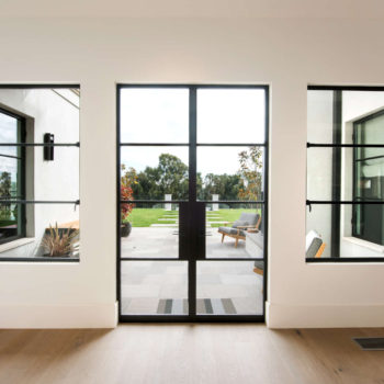 Contemporary home interior illuminated by Euroline Steel Casement Windows and doors, featuring floor-to-ceiling black steel-framed glass panels that open to a lush outdoor patio, seamlessly blending the indoor living space with the natural surroundings.