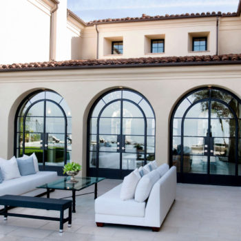 Luxurious patio setting featuring Euroline Steel Swing Doors with arched design and black frames, enhancing the Mediterranean-style architecture of the home. The spacious terrace is furnished with modern outdoor sofas, providing a comfortable area to enjoy the expansive views of the waterfront.