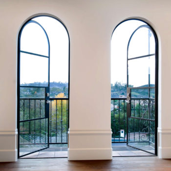 Elegant interior featuring Euroline Steel Swing Doors with arched tops and intricate black frames, set against a clean white wall. These doors open to a balcony, offering a serene view of the lush landscape beyond.