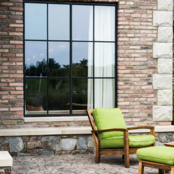 Relaxing outdoor seating area enhanced by Euroline Steel Curtain Wall Windows, featuring a large, black steel-framed window set against a rustic brick and stone wall. The space is accented with bright green lounge chairs, offering a cozy spot to enjoy the natural surroundings.