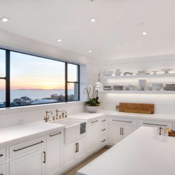Bright and modern kitchen featuring Euroline Steel Casement Windows, with wide glass panels framed in sleek black steel that offer stunning sunset views over the ocean. The kitchen is fitted with white cabinetry and a spacious center island, providing a clean and inviting atmosphere.